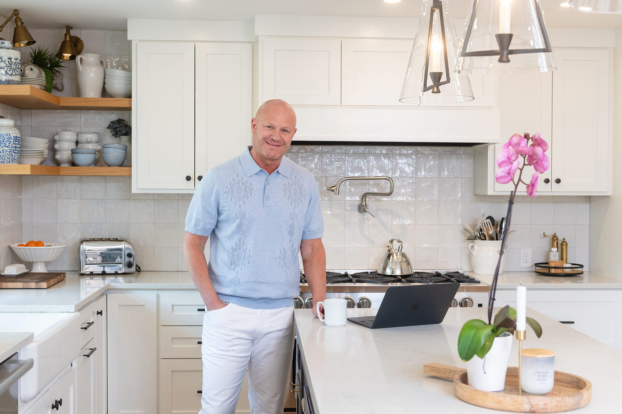 man in a light blue shirt and white pants standing in a white kitchen with his coffee